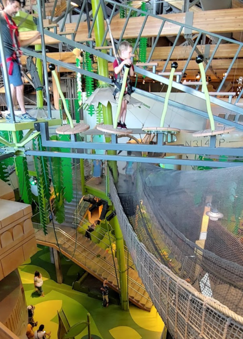 A child on a high ropes course, with adults and other children below in a vibrant indoor play area.