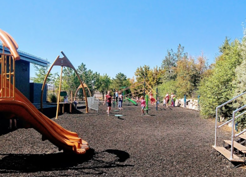 A playground with a slide and swings, children playing on a sunny day surrounded by trees and a blue sky.