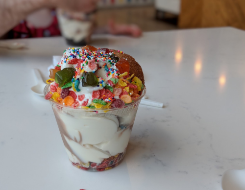 A colorful ice cream sundae topped with sprinkles, candy, and a cherry in a clear cup on a marble table.