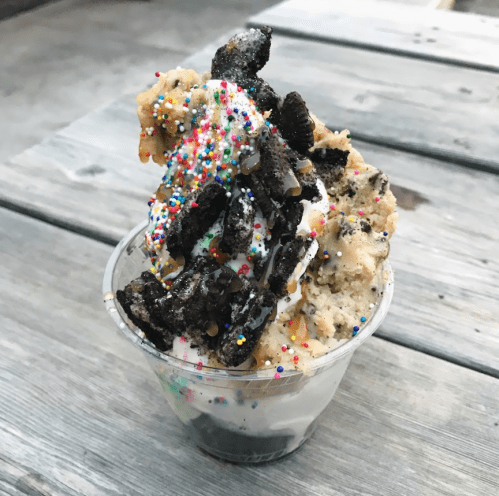 A cup of ice cream topped with cookie crumbles, crushed Oreos, and colorful sprinkles on a wooden table.