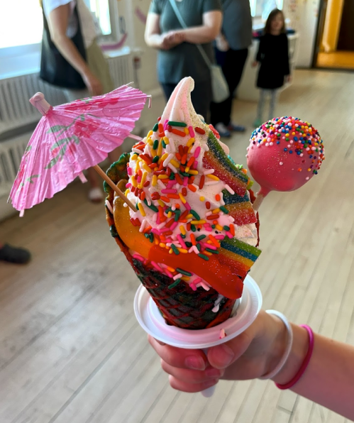 A colorful ice cream cone topped with sprinkles, a mini umbrella, and a candy pop, held by a person.