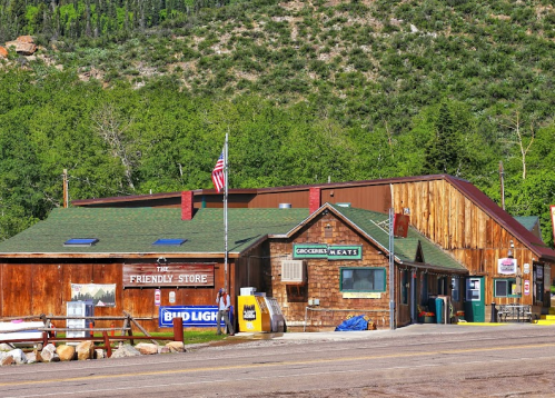 A rustic store with wooden siding, an American flag, and a sign for groceries and meats, set against a green hillside.