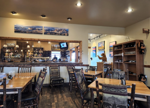 Cozy restaurant interior with wooden tables, a bar area, and mountain-themed decor. A bear statue is visible in the background.