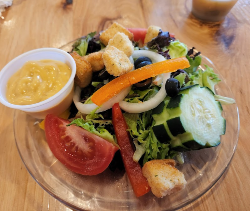 A fresh salad with mixed greens, cucumbers, tomatoes, peppers, olives, croutons, and a side of dressing.