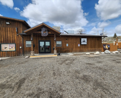 A rustic wooden building with a stone entrance, surrounded by gravel and a cloudy sky.