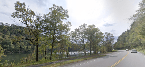 A scenic view of a winding road alongside a calm lake, framed by trees and rolling hills under a cloudy sky.