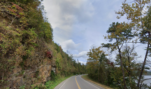 A winding road alongside a rocky cliff and trees, with a body of water visible in the distance under a cloudy sky.
