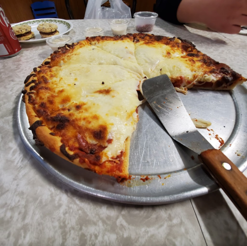 A partially sliced cheese pizza on a metal tray, with a pizza cutter and cookies in the background.