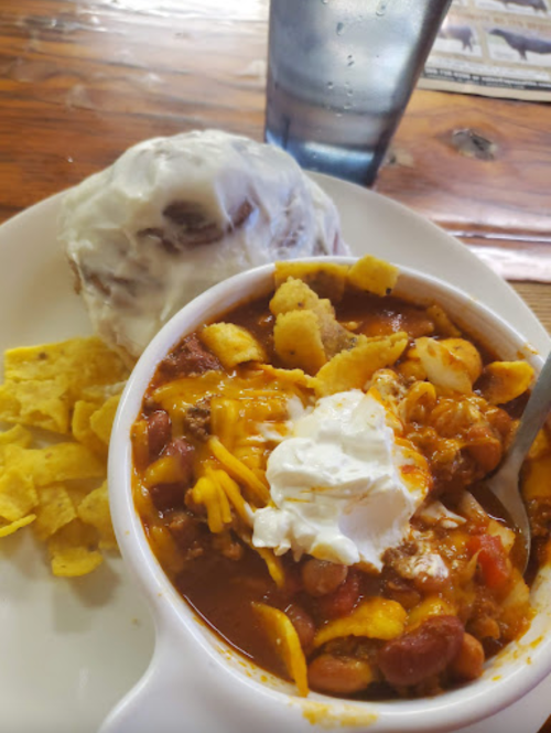 A bowl of chili topped with cheese and sour cream, served with tortilla chips and a cinnamon roll on the side.