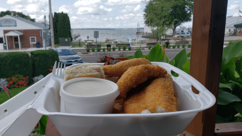 A hand holds a takeout container with fried fish, a roll, and dipping sauce, with a lake view in the background.