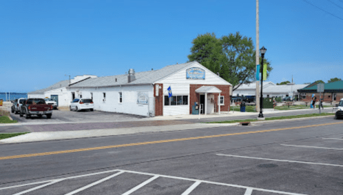 A white building with a brick facade, parked cars, and a clear blue sky in a small town near the water.