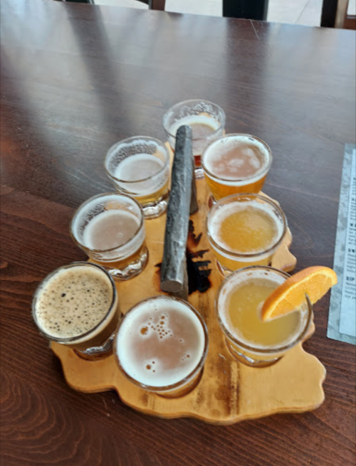 A wooden flight board with eight small glasses of different beers, featuring a slice of orange on one glass.