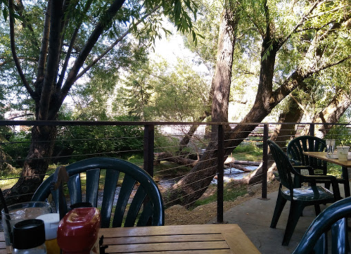 A serene outdoor dining area with wooden tables, chairs, and a view of trees and a stream in the background.