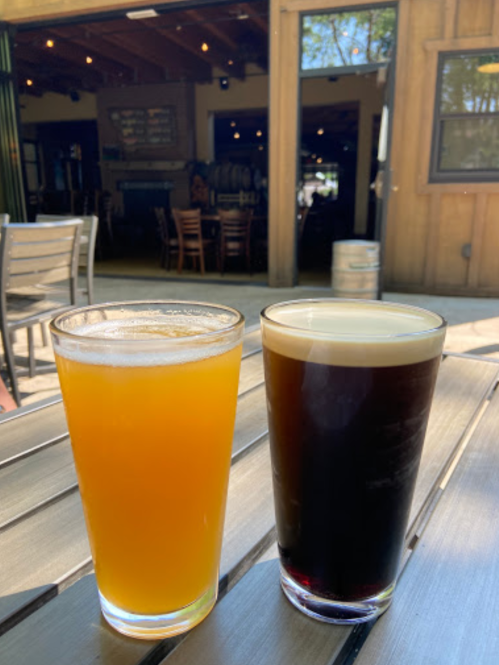 Two glasses of beer sit on a wooden table, one pale and frothy, the other dark with a creamy head, in a sunny outdoor setting.