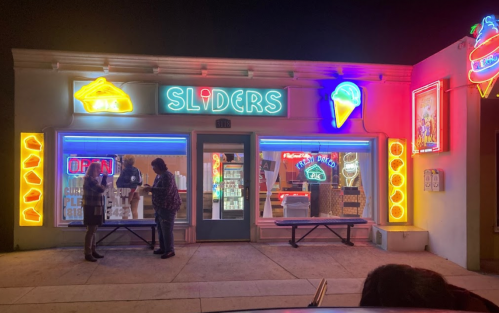 A brightly lit diner named "Sliders" with neon signs, featuring two people chatting outside at night.