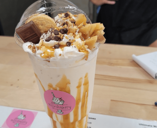 A tall milkshake topped with whipped cream, chocolate, cookies, and caramel drizzle in a clear cup.