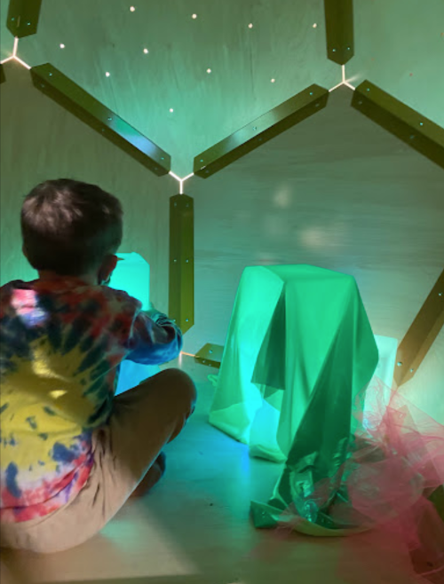 A child in a tie-dye shirt interacts with glowing objects in a colorful, illuminated play space.
