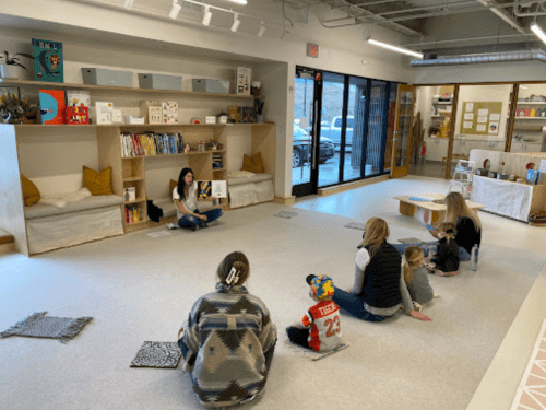 A group of children and adults sit on the floor, listening to a storyteller in a cozy, bright space.