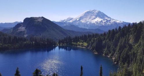 A serene mountain landscape featuring a blue lake, lush forests, and a snow-capped peak in the background.