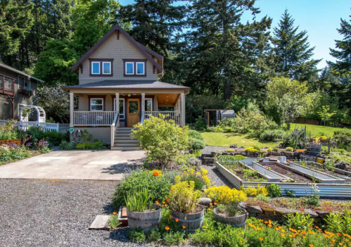 A charming house with a porch surrounded by a vibrant garden and trees under a clear blue sky.