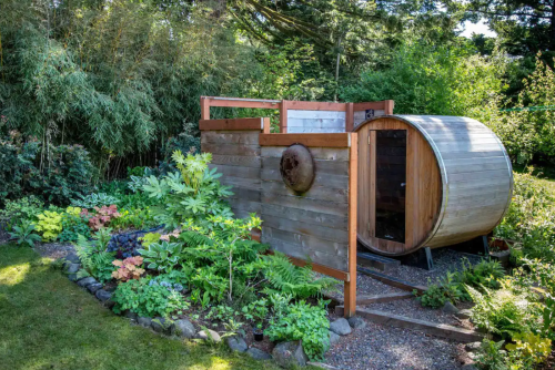 A wooden sauna barrel surrounded by lush greenery and colorful plants in a serene garden setting.
