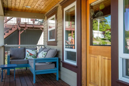 A cozy porch with a blue couch, decorative pillows, and a wooden door, surrounded by greenery and a staircase in the background.