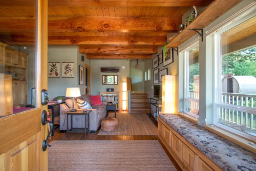 Cozy living room with wooden beams, a sofa, and natural light from large windows, featuring a warm, inviting atmosphere.