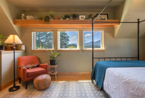 Cozy bedroom with a bed, armchair, plants, and large windows showcasing a mountain view. Warm, inviting atmosphere.