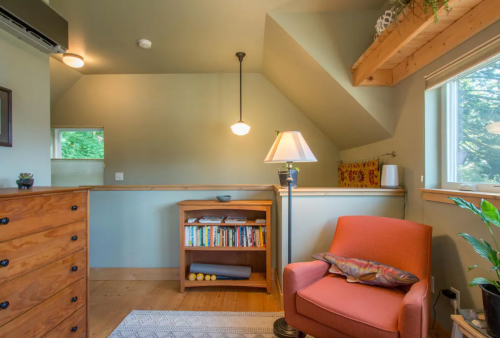 Cozy room with sloped ceiling, wooden furniture, a lamp, bookshelves, and an orange armchair by a window.