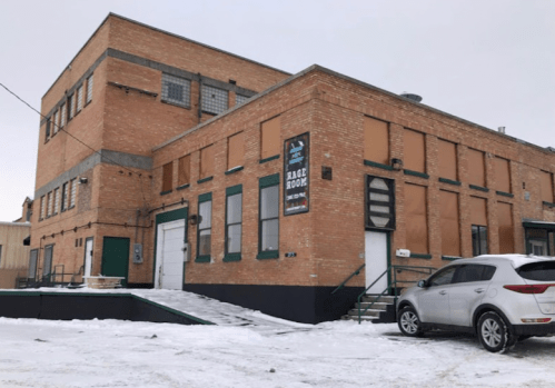 A brick building with boarded windows and a sign reading "Rage Room," surrounded by snow and a parked car.