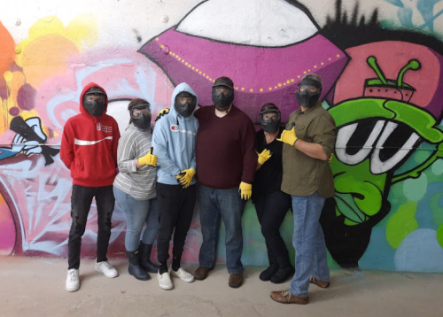 A group of six people wearing masks and gloves poses in front of a colorful graffiti wall.