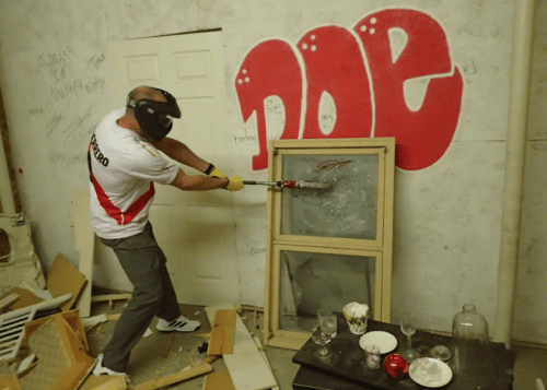 A person in protective gear swings a bat at a shattered window in a messy room with graffiti on the wall.