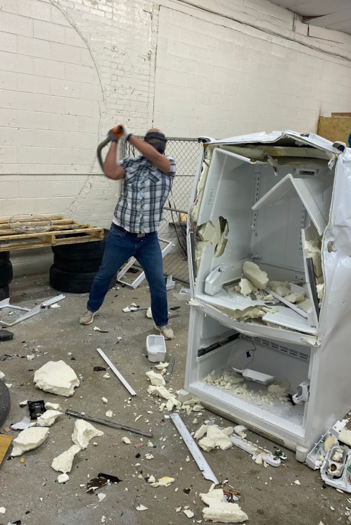 A person in a plaid shirt swings a sledgehammer at a broken refrigerator in a messy workshop.