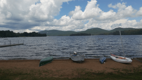 A serene lake scene with boats on the shore, surrounded by mountains and a cloudy sky.
