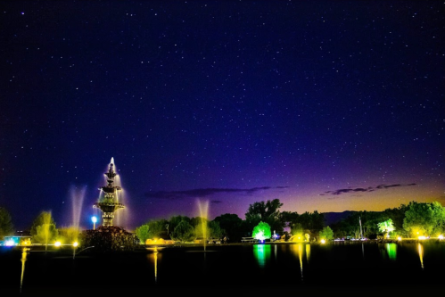 A serene night scene featuring a fountain surrounded by trees, under a starry sky with vibrant colors reflecting on the water.