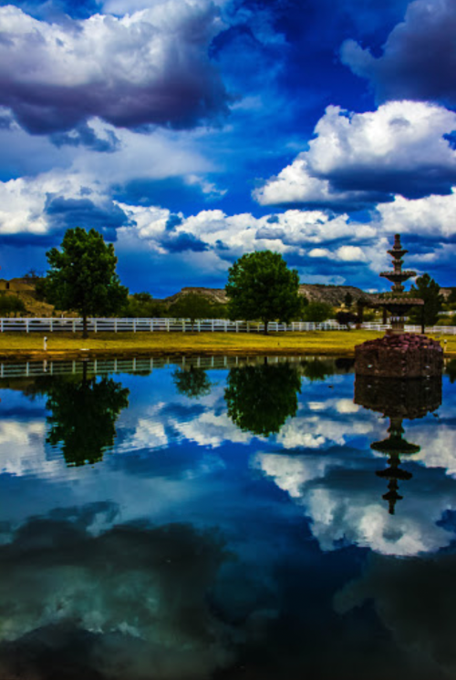 A serene pond reflects fluffy clouds and trees under a vibrant blue sky, with a fountain in the foreground.