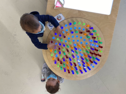 Two children play with colorful translucent pieces on a circular table, exploring patterns and colors.