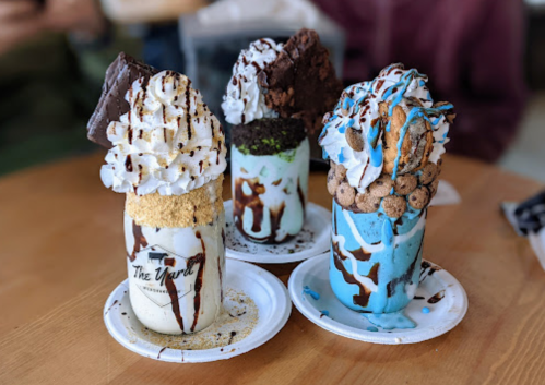 Three colorful, over-the-top milkshakes topped with whipped cream, cookies, and chocolate drizzles on white plates.