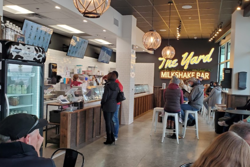 Interior of a milkshake bar with a counter, customers, and a large neon sign reading "The Yard Milkshake Bar."