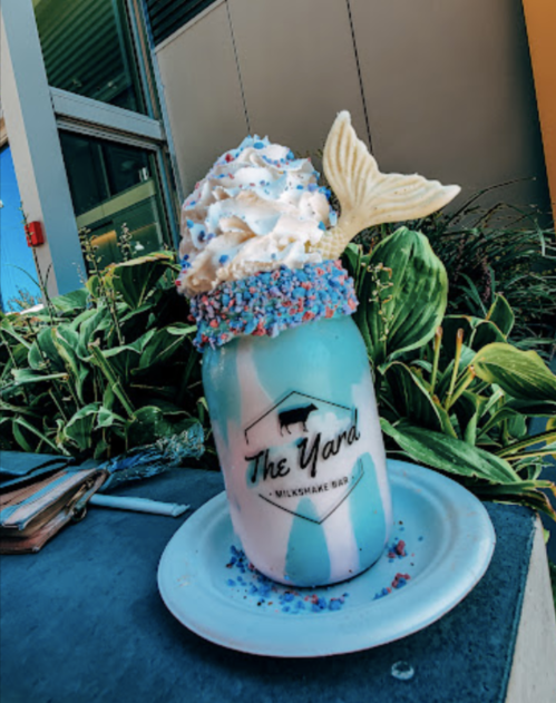 A colorful milkshake topped with whipped cream, sprinkles, and a white chocolate mermaid tail, placed on a blue plate.