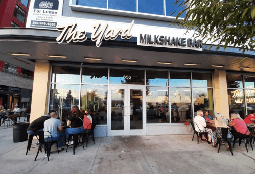 Exterior of The Yard Milkshake Bar, featuring outdoor seating and large windows, with people enjoying milkshakes.