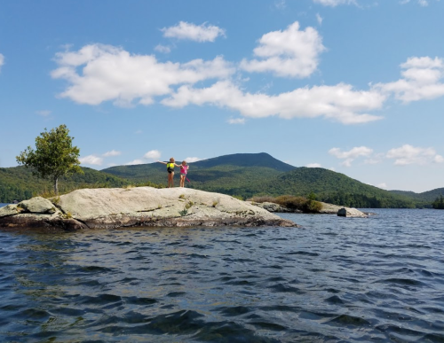 Two people stand on a rocky outcrop by a lake, surrounded by green mountains and a blue sky with fluffy clouds.