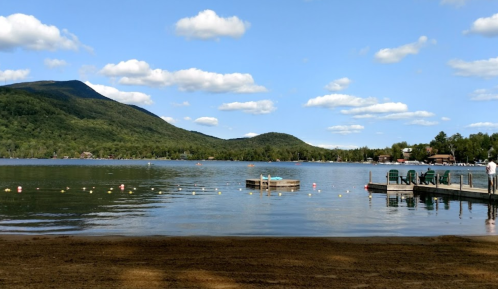 A serene lake surrounded by mountains, with a dock and a small floating platform under a blue sky with fluffy clouds.