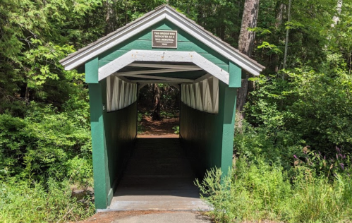 A green covered bridge surrounded by trees, leading into a wooded area. A sign is visible above the entrance.