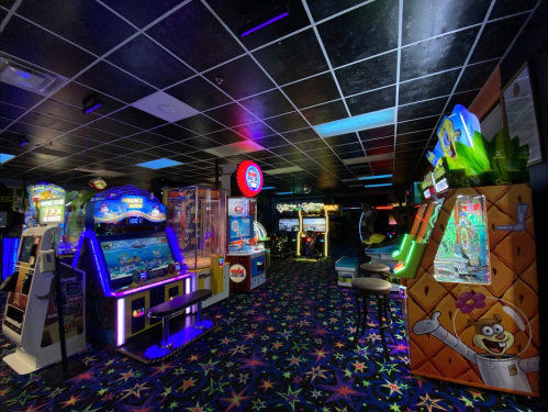 A brightly lit arcade with various gaming machines and colorful carpet under a black ceiling.
