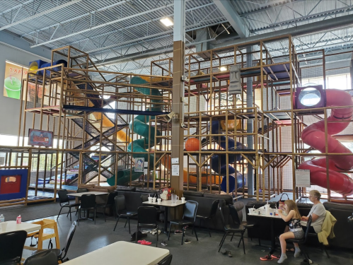 Indoor play structure with slides and climbing areas, surrounded by tables and chairs in a spacious recreational area.