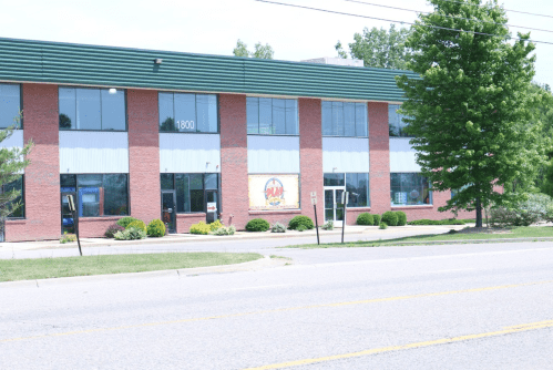 A brick building with large windows and a green roof, featuring a sign and landscaping along a street.