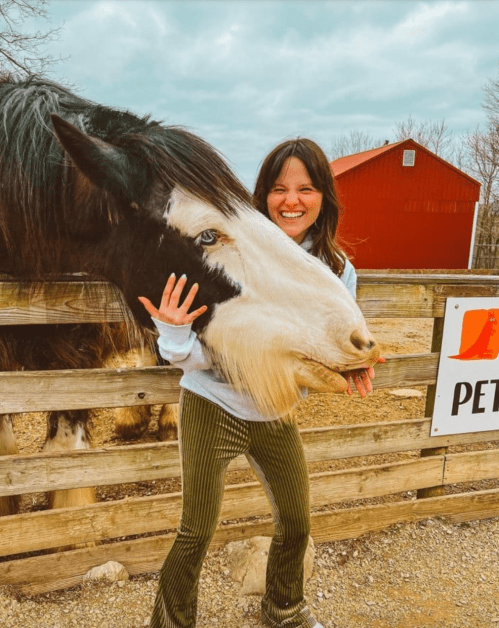 A woman smiles while playfully interacting with a large horse at a petting farm, with a red barn in the background.