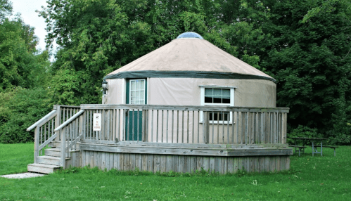 A round yurt with a wooden deck, surrounded by green grass and trees.