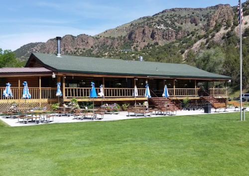 A wooden lodge with a green roof, surrounded by mountains, featuring outdoor seating and blue umbrellas.